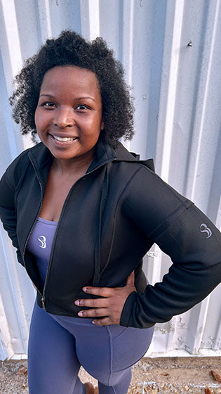 Woman wearing a black jacket and purple leggings standing against a corrugated metal background